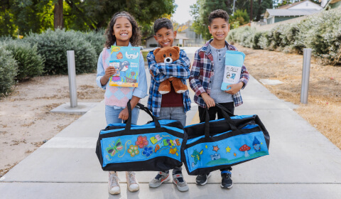 Three smiling young children with duffel bags and toys provided by Foster Love